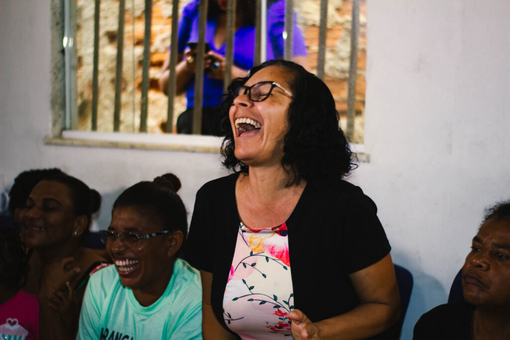 À esquerda, duas mulheres negras sentadas sorrindo. Ao meio, uma mulher negra de cabelos curtos ri. À direita, uma mulher negra sentada com rosto sério. Atrás, uma janela gradeada.