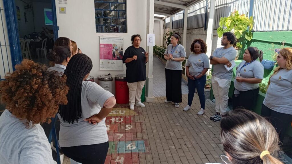 Um grupo composto por mulheres e homens se reúne em pé, formando um círculo, e escutam o educador territorial em uma atividade do percurso formativo.