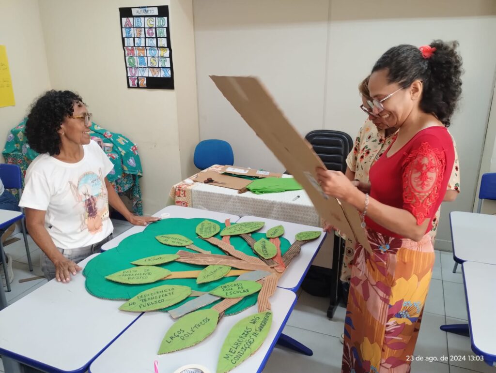 Três mulheres montam uma árvore de papelão, com folhas penduradas nos galhos. Em cada folha contém, escritos à mão, os desafios locais de Belém, Pará, mapeados pelos participantes do percurso formativo.