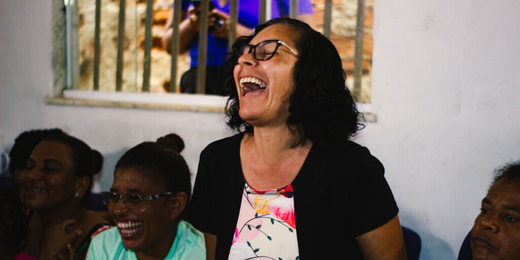 À esquerda, duas mulheres negras sentadas sorrindo. Ao meio, uma mulher negra de cabelos curtos ri. À direita, uma mulher negra sentada com rosto sério. Atrás, uma janela gradeada.